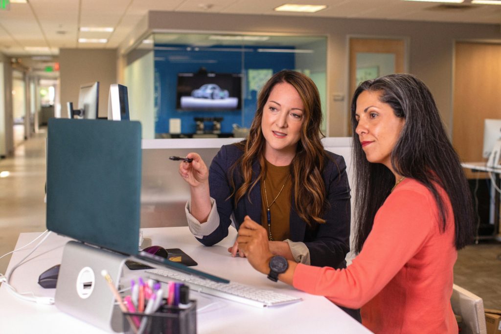 Two women seated at an office desk looking at a computer. 